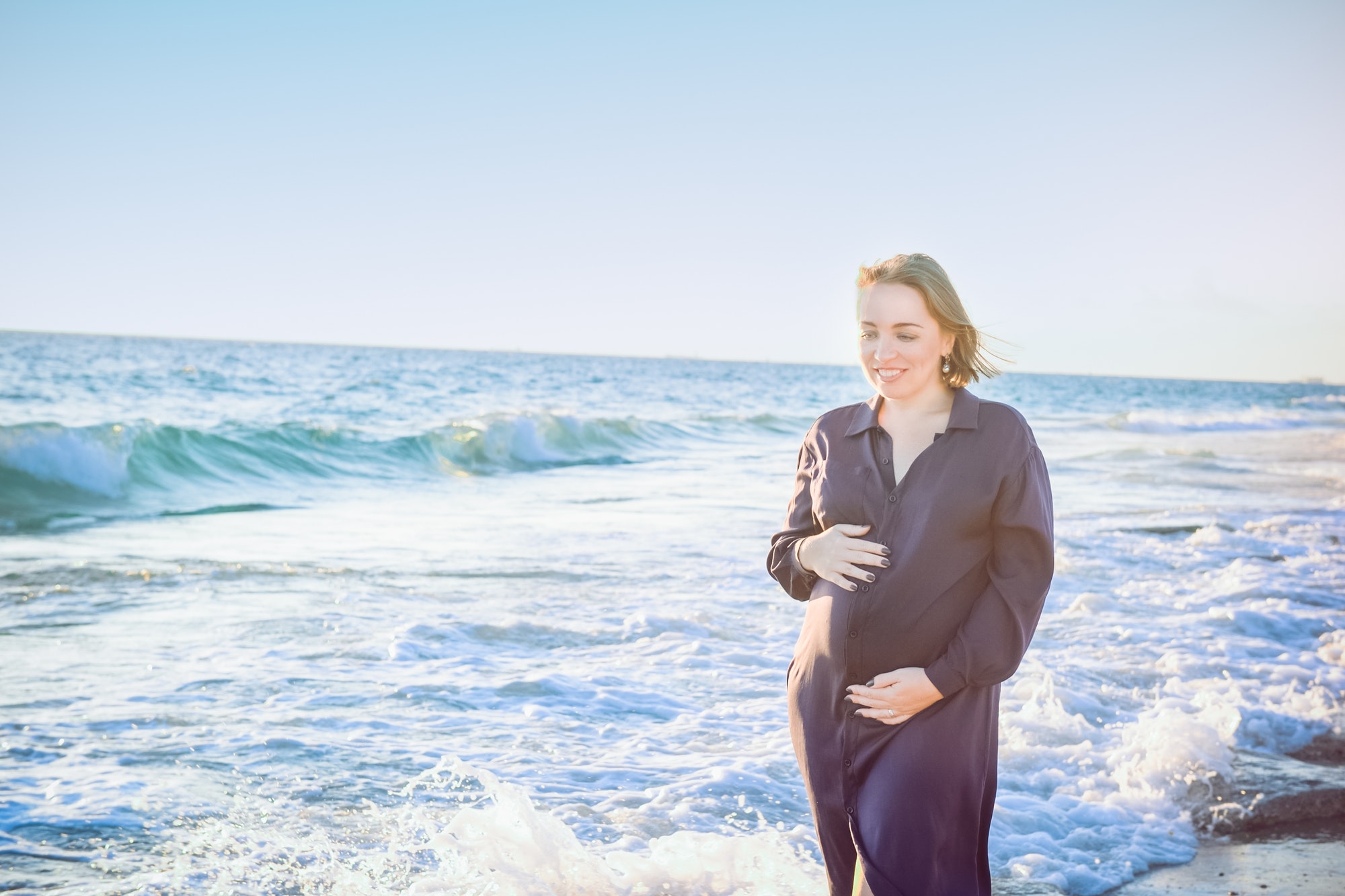 pregnant woman in dark blue dress touching her belly with love and care walking and on the beach.