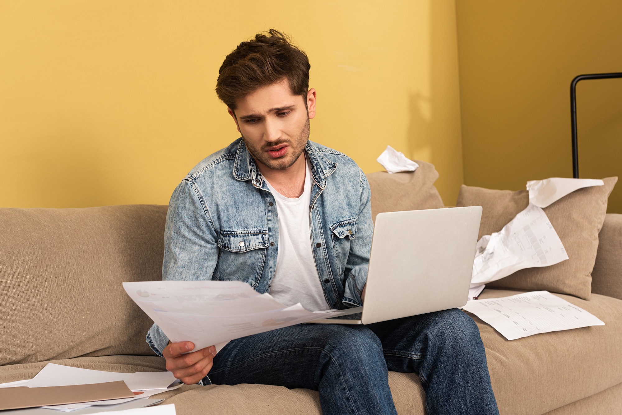 Sad man working with papers and laptop on couch at home